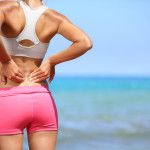 Back pain. Athletic woman in pink sportswear standing at the seaside rubbing the muscles of her lower back, cropped torso portrait.