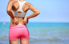 Back pain. Athletic woman in pink sportswear standing at the seaside rubbing the muscles of her lower back, cropped torso portrait.
